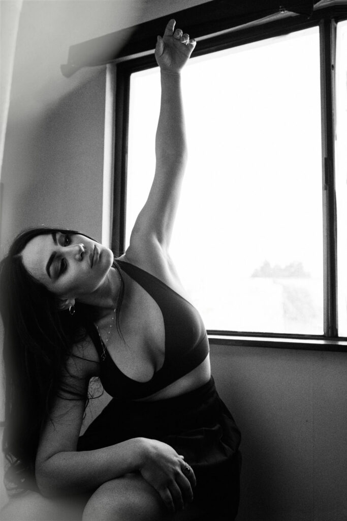 Valeria Cisneros poses elegantly by a window stretching her arm upwards wearing a deep V-neck top and a skirt.